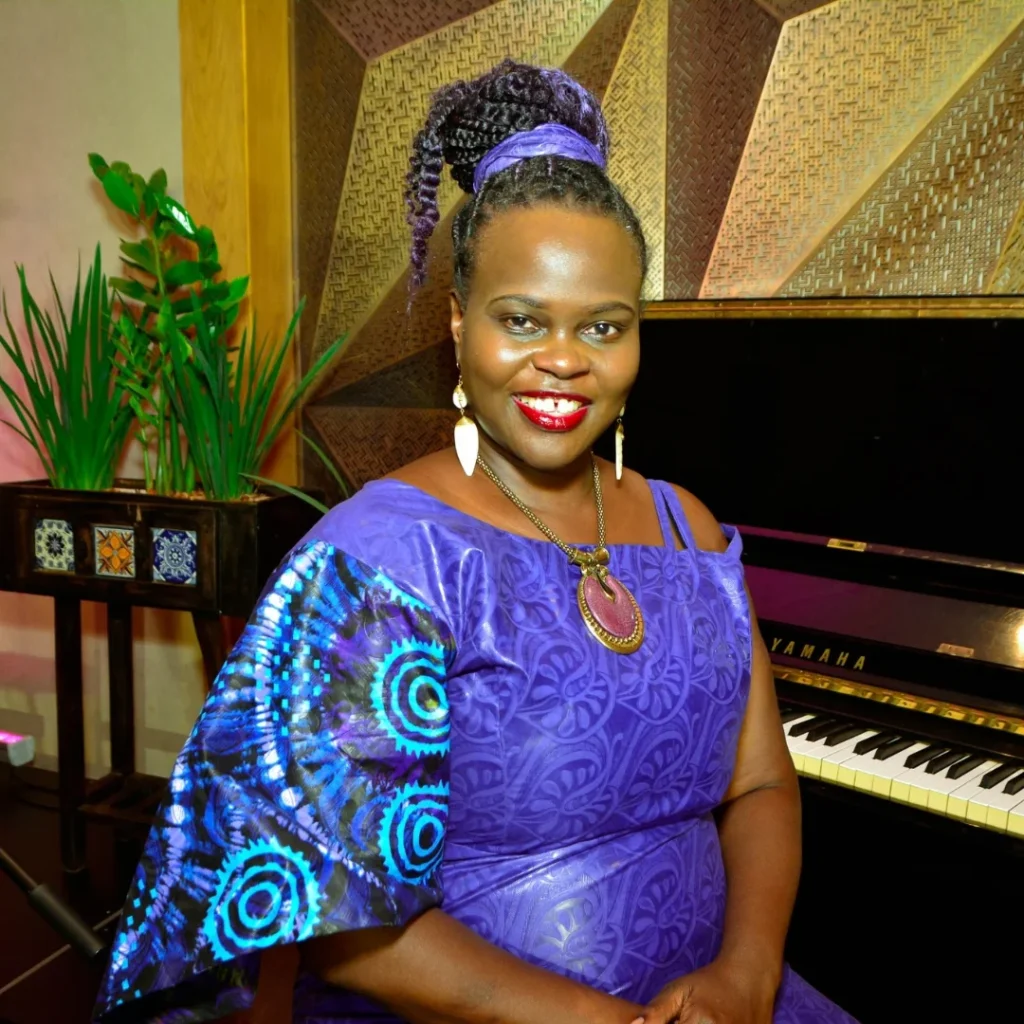 Beverley in a purple African dress seated next to a piano.